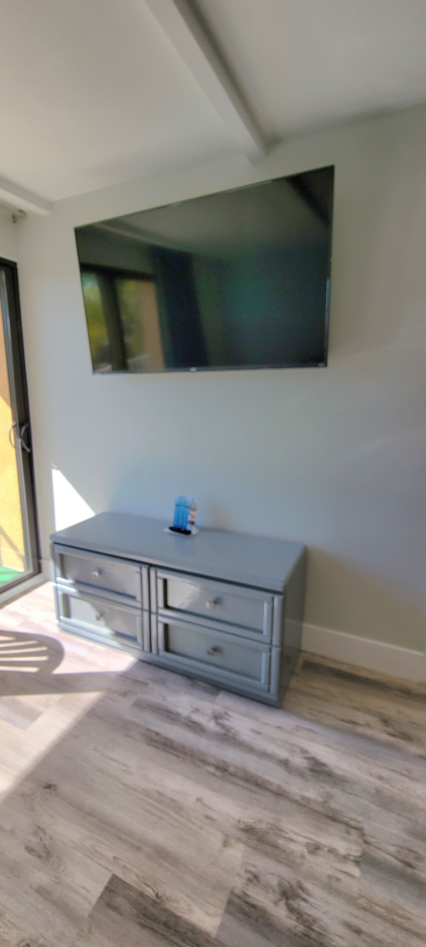 A room with a large flat-screen TV mounted on the wall, a grey cabinet with drawers, and a wooden floor.