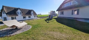 A courtyard with two white buildings, one with a thatched roof and the other with a flat roof. There is a wooden bench and a small play structure in the center.