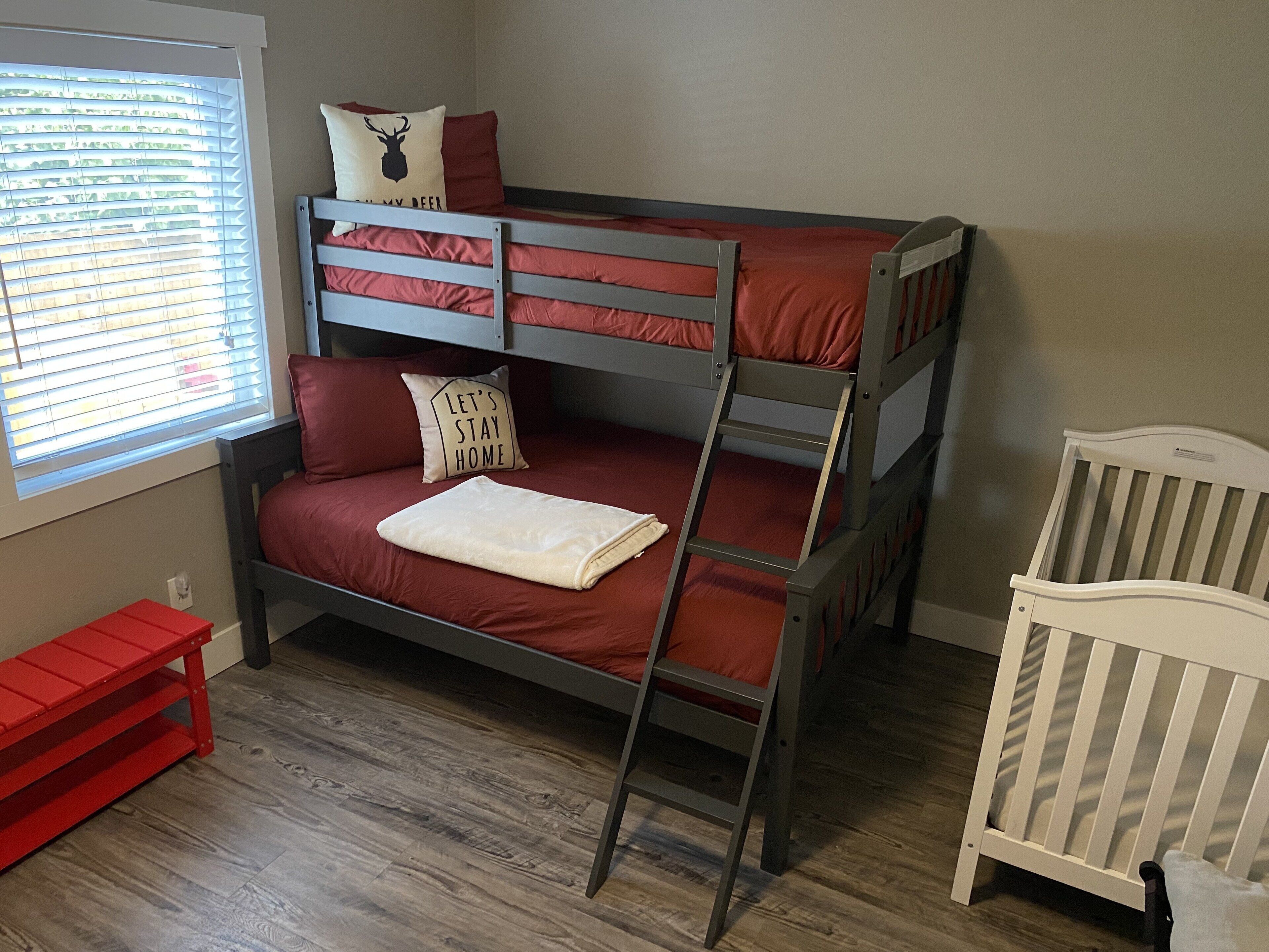 A bunk bed with red bedding and a ladder, a white crib, and a red bench.