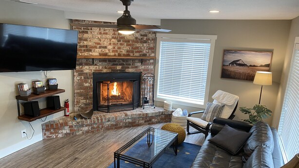 A living room with a brick fireplace, a flat-screen TV mounted above it, a sofa, a coffee table, and a window with blinds. - Spacious 5BR with Sauna & AC, Fenced Yard,Fast WiFi, Near Willamette Pass (Oakridge)