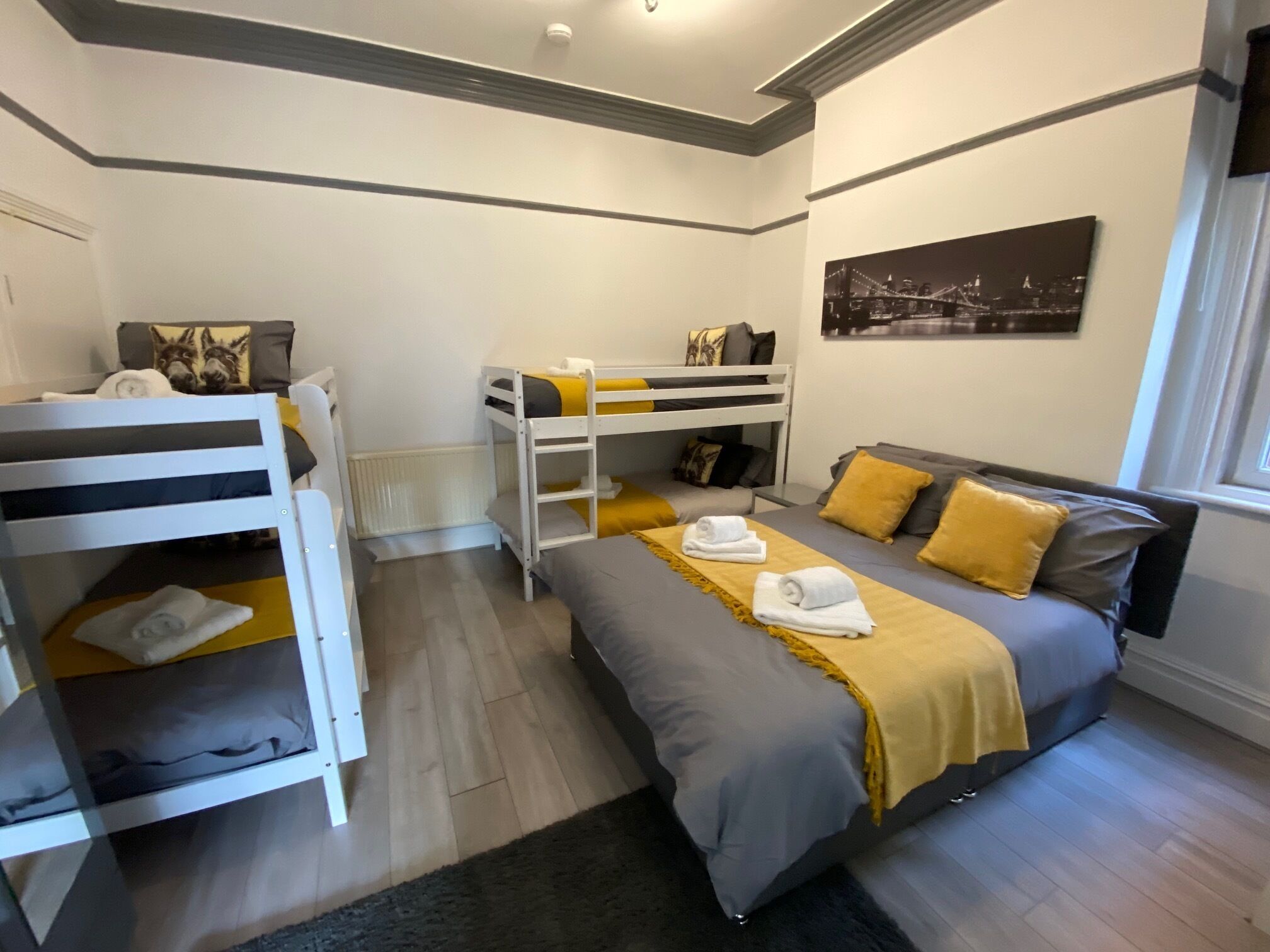 Un dormitorio con literas, una cama con manta amarilla y un cuadro de paisaje urbano en la pared.