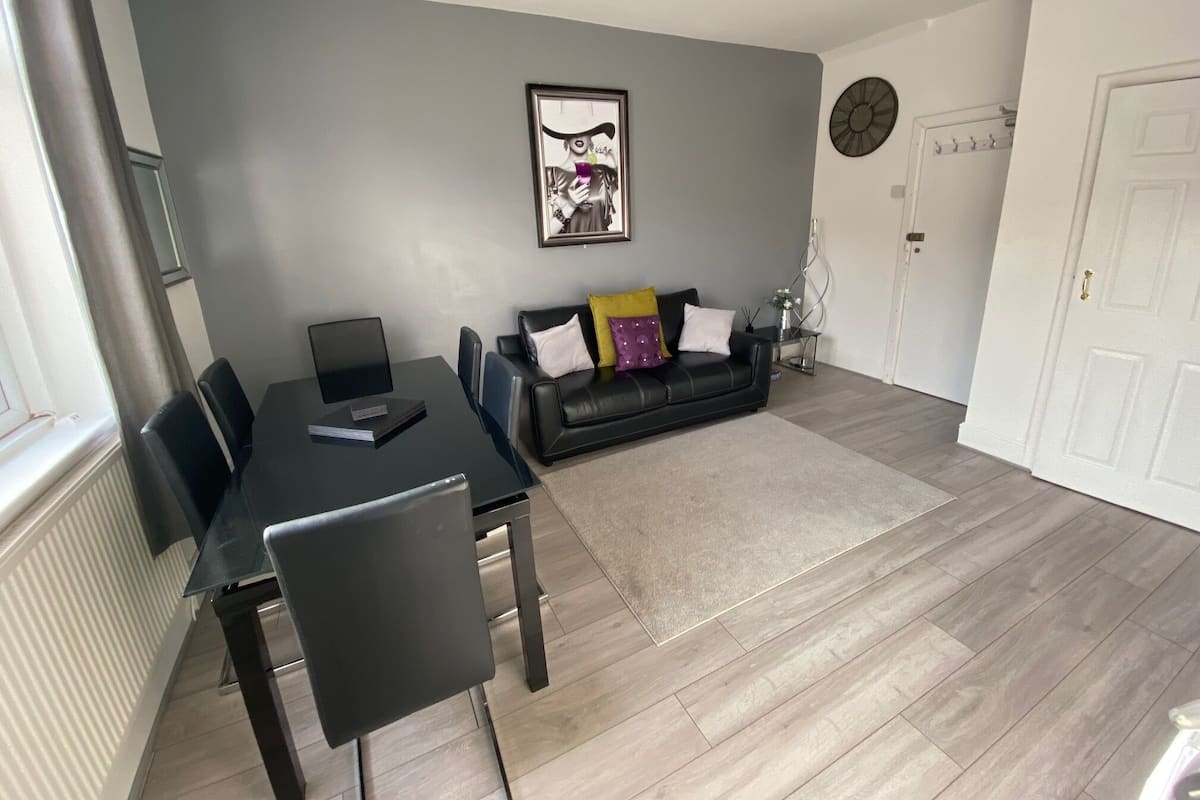 A modern living room with a black leather sofa, a dining table with chairs, and a grey wall with a framed picture.