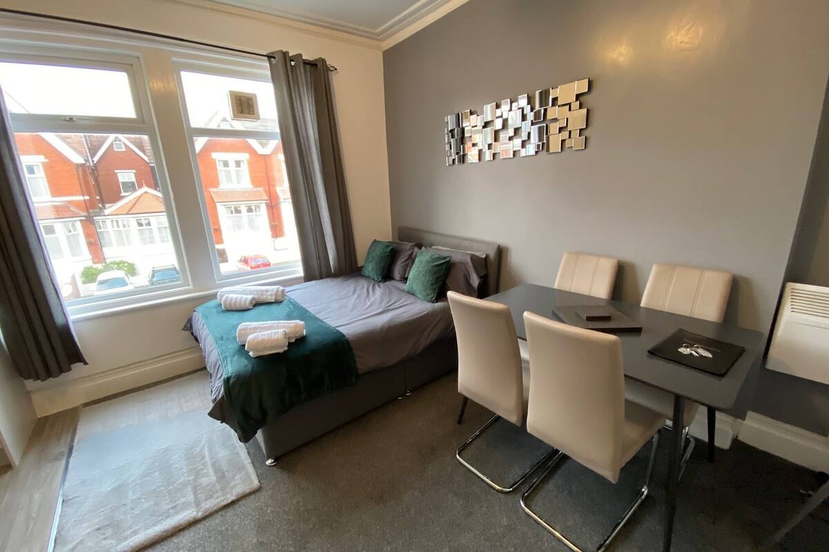 A hotel room with a bed, a desk, and a chair. There is a window with curtains, a wall-mounted artwork, and a view of buildings outside.