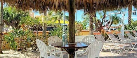 A thatched umbrella table with white plastic chairs, set on a paved patio with palm trees in the background.