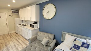 A compact room with a kitchenette, a grey sofa, a bed with patterned bedding, and a wall clock.
