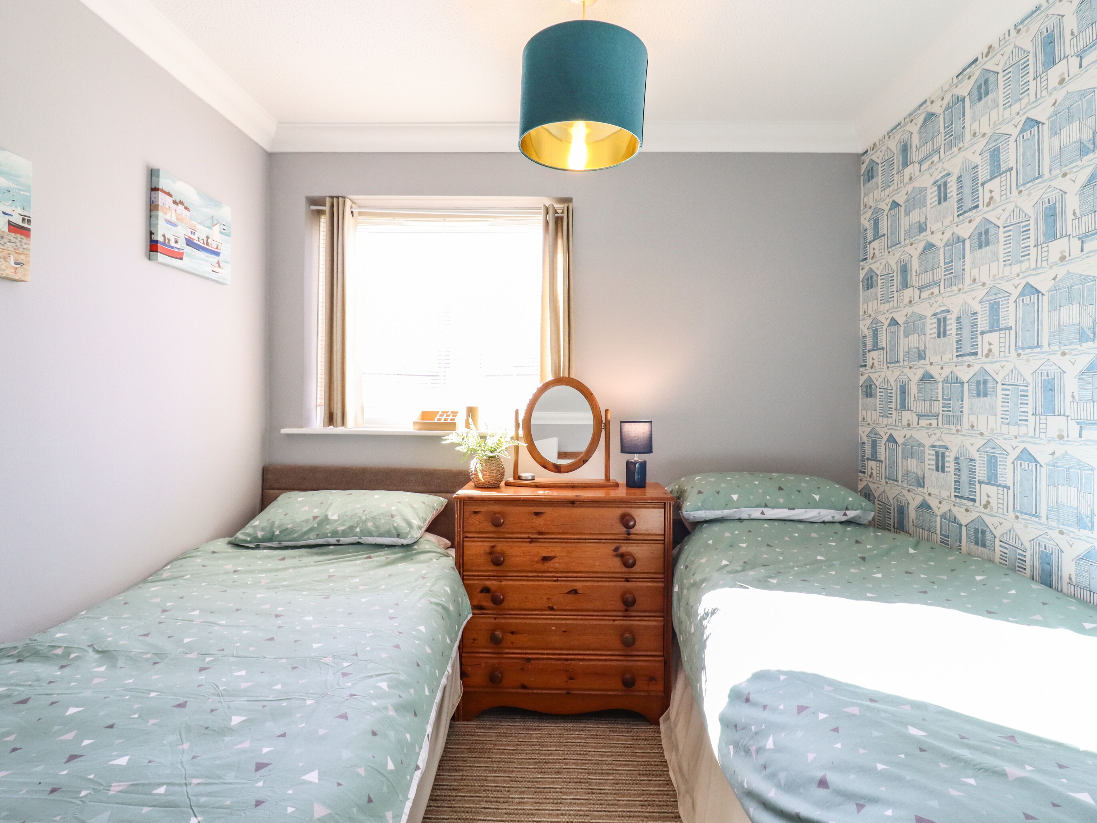 Une chambre avec deux lits simples, une commode, un miroir, une fenêtre et un mur décoré d’un motif.