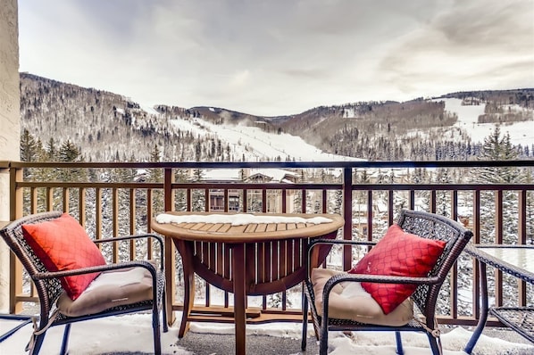 A balcony with wicker chairs and a table, overlooking a snowy mountain landscape.