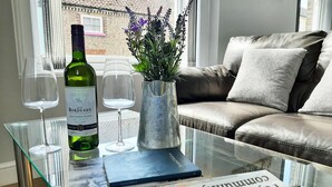 A living room with a glass coffee table holding a bottle of wine, a vase with flowers, a remote control, and a book. - Malton Central Apartments (Malton)