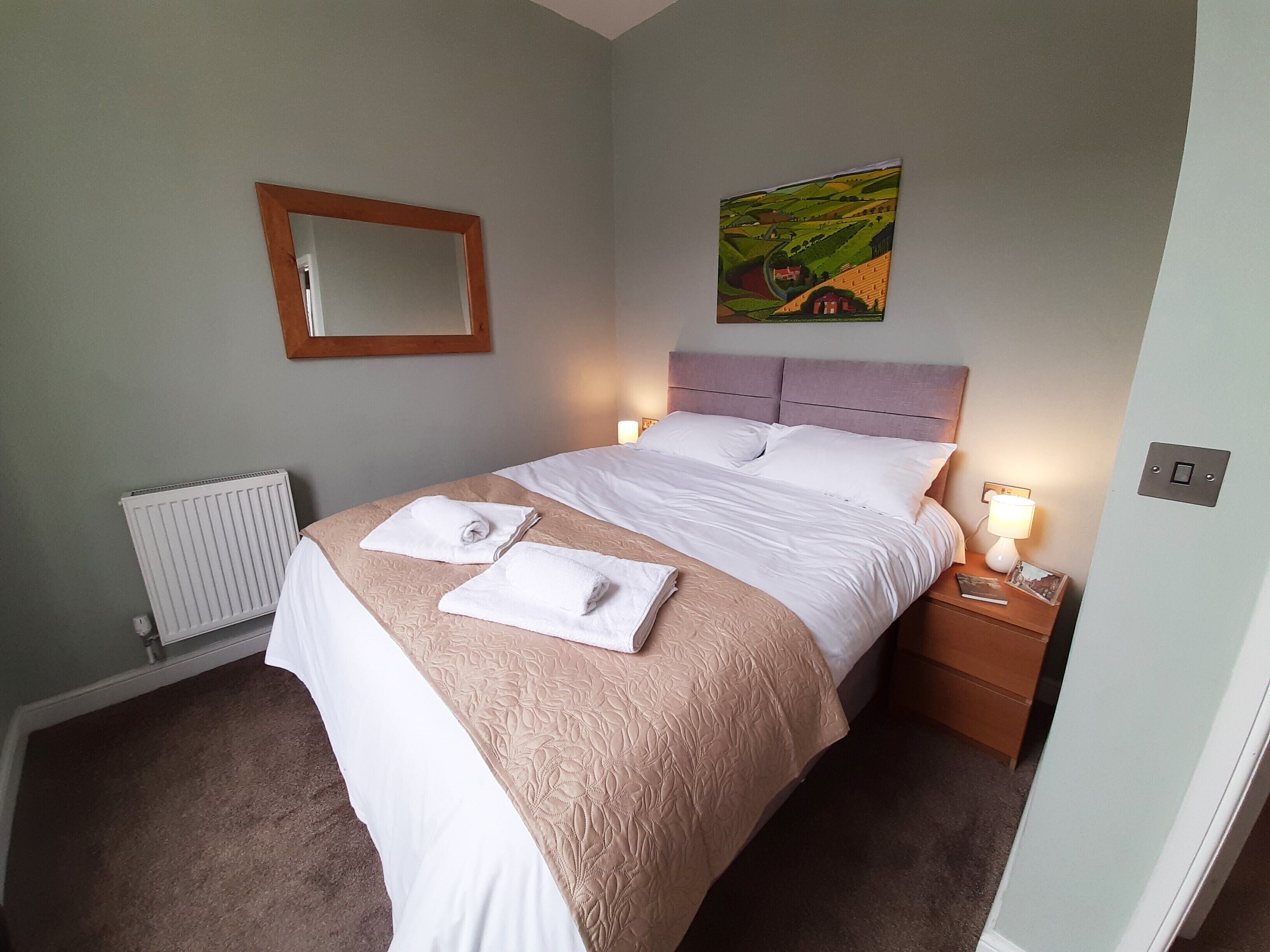 A neatly made bed with white linens and a beige bedspread, a bedside table with a lamp, a framed picture on the wall, and a radiator on the floor.