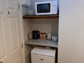Una cocina compacta con microondas en un estante, cafetera, tazas y un refrigerador pequeño.