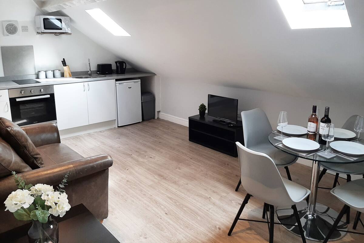 A modern kitchen with a built-in microwave, oven, and dishwasher. There is a dining area with a table set for four, a sofa, and a vase with flowers.