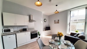 A modern kitchen with white cabinets, a microwave, and a sink. There is a dining area with a glass table, chairs, and a vase of yellow flowers. A sofa and a TV are visible in the background. - Malton Central Apartments (Malton)