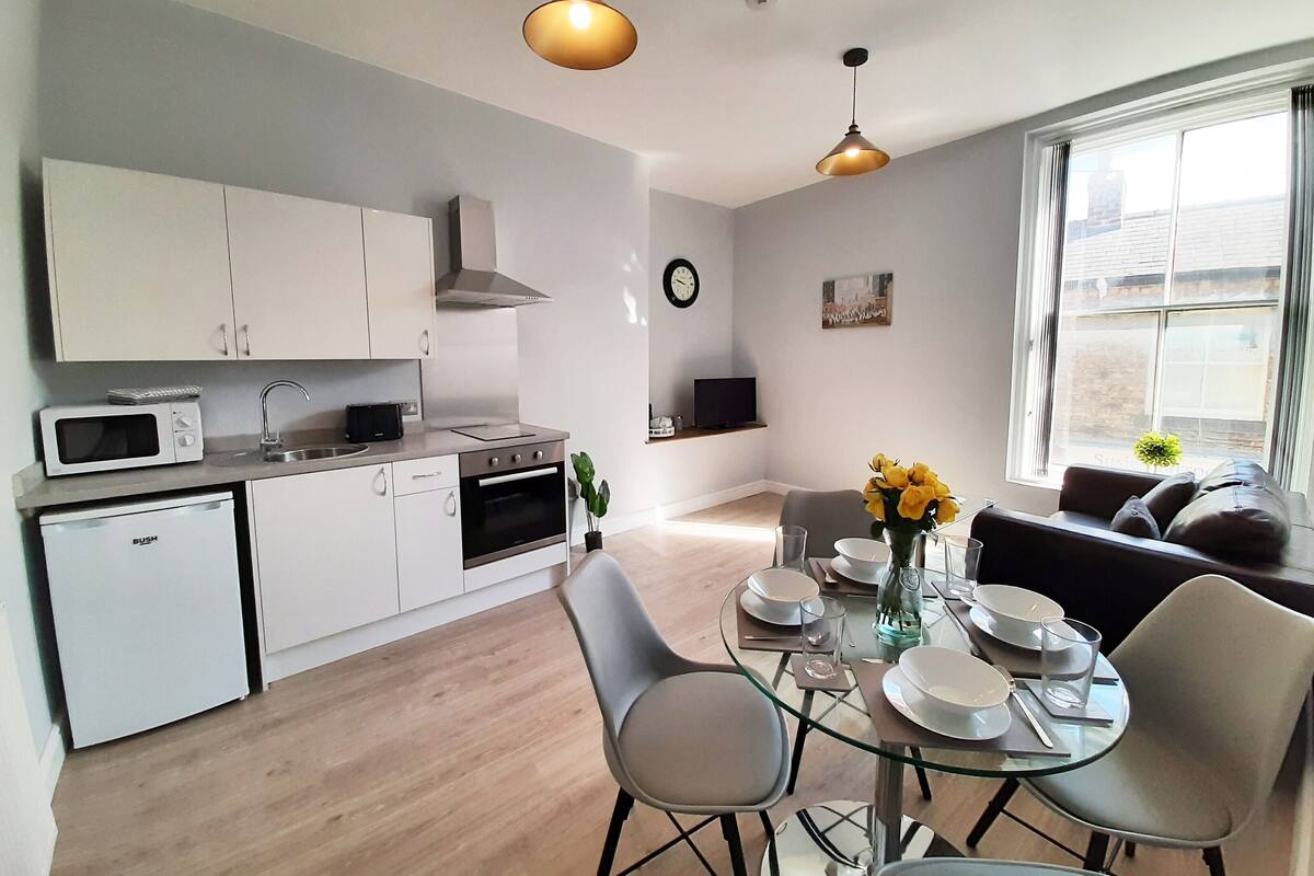 A modern kitchen with white cabinets, a microwave, and a sink. There is a dining area with a glass table, chairs, and a vase of yellow flowers. A sofa and a TV are visible in the background.