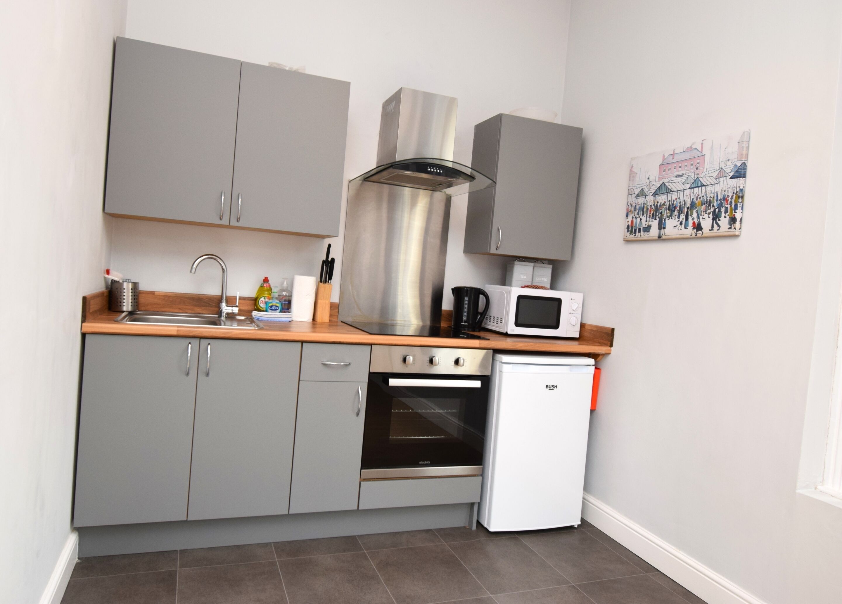 A compact kitchen with grey cabinets, a stainless steel range hood, a microwave, a small oven, and a mini fridge.