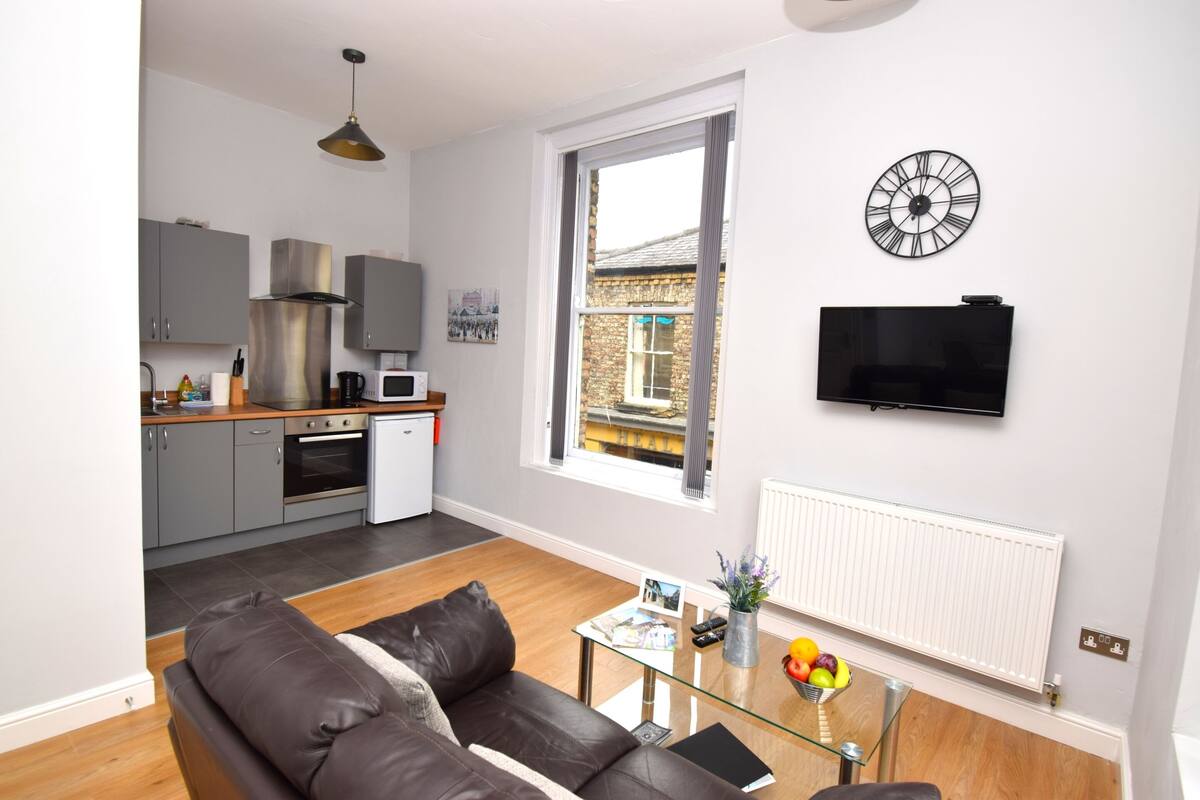 A modern living room with a sofa, a glass coffee table, a TV, and a kitchen in the background.