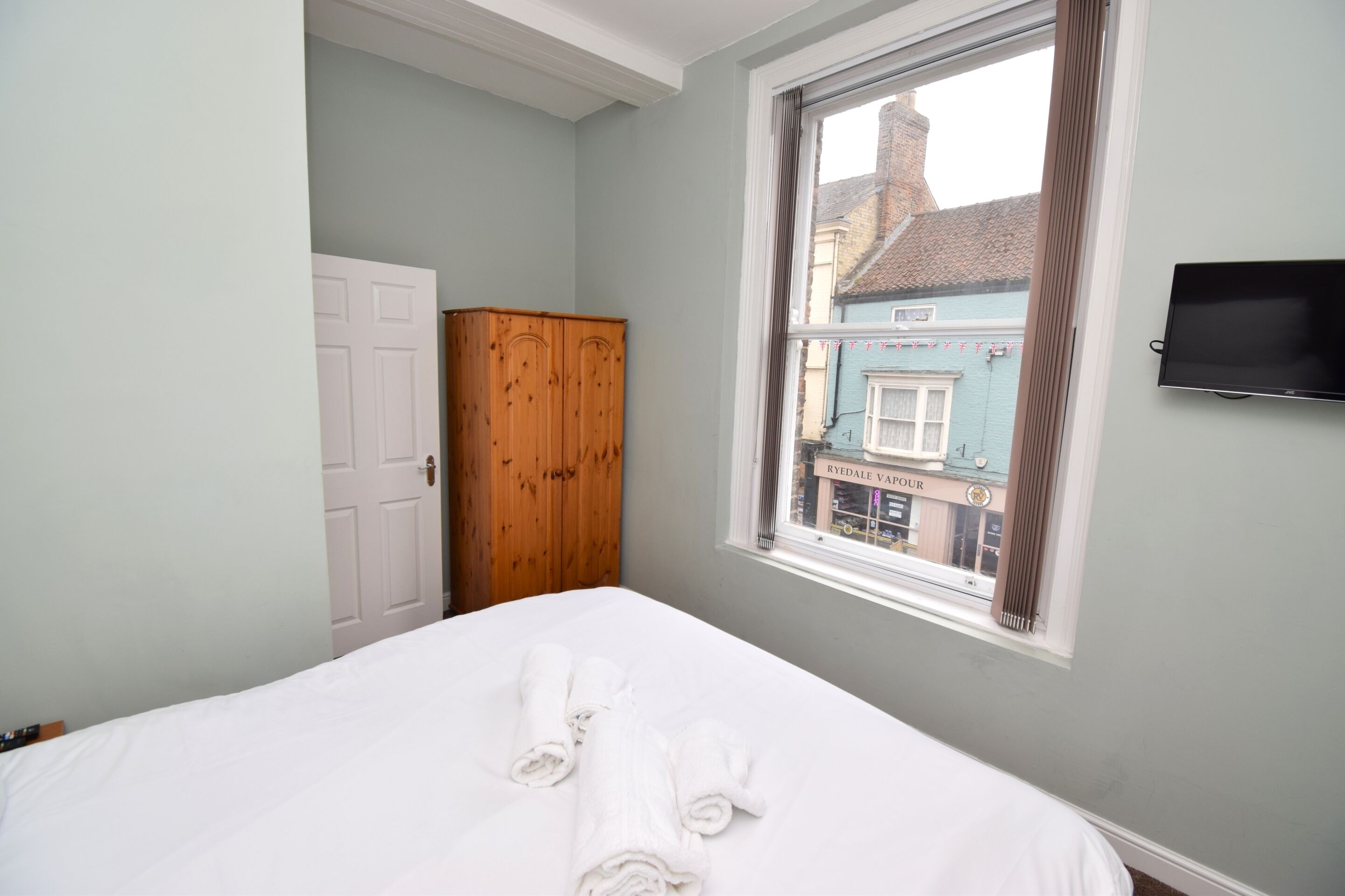 A bedroom with a bed, a wardrobe, a window with a view of a building, and a TV mounted on the wall.