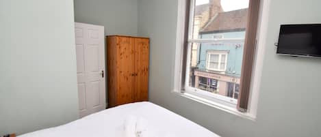 A bedroom with a bed, a wardrobe, a window with a view of a building, and a TV mounted on the wall.