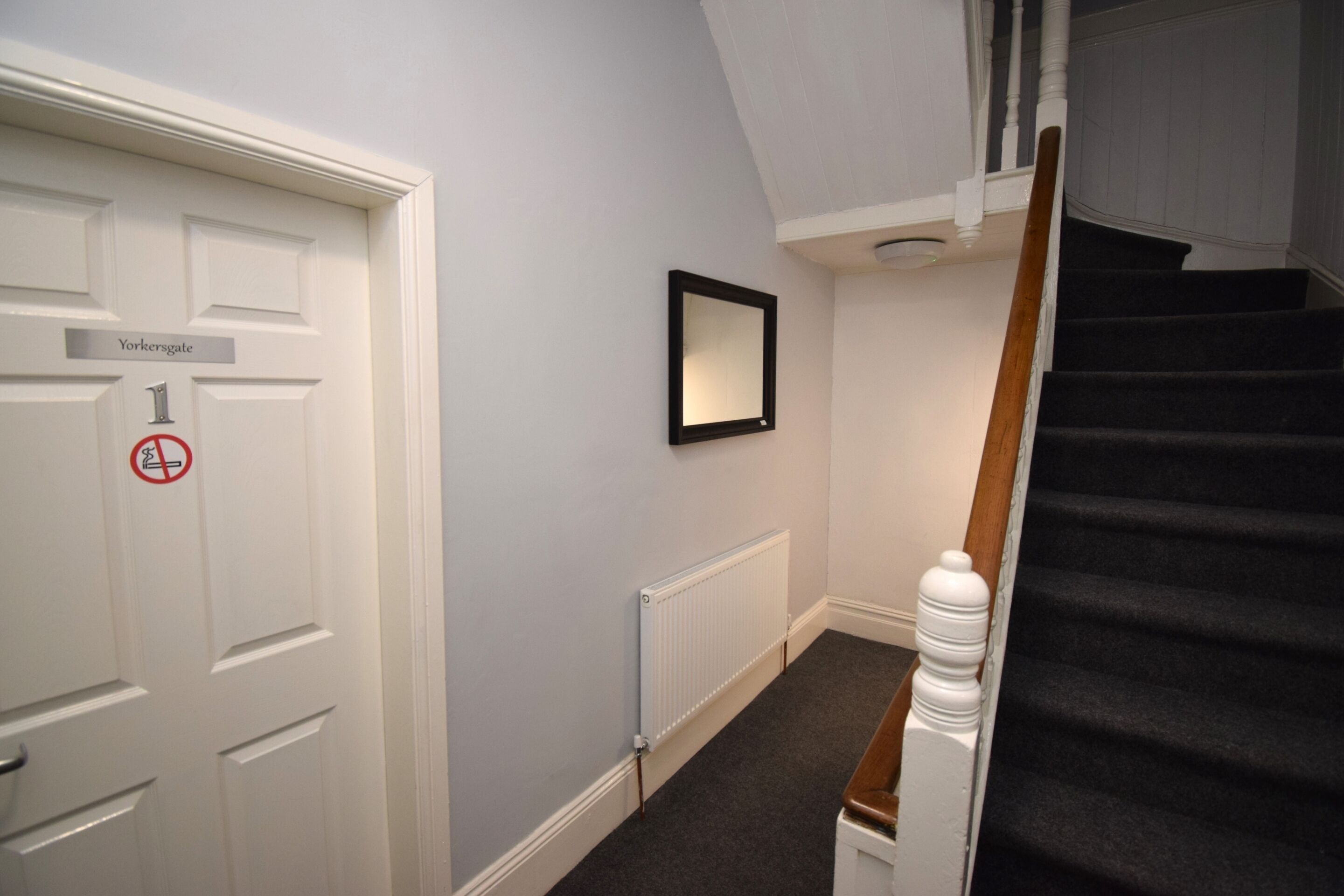 Un couloir avec une porte blanche portant l’inscription « Yorkersgate 1 », un miroir encadré de noir accroché au mur, un radiateur et un escalier avec une rampe en bois.