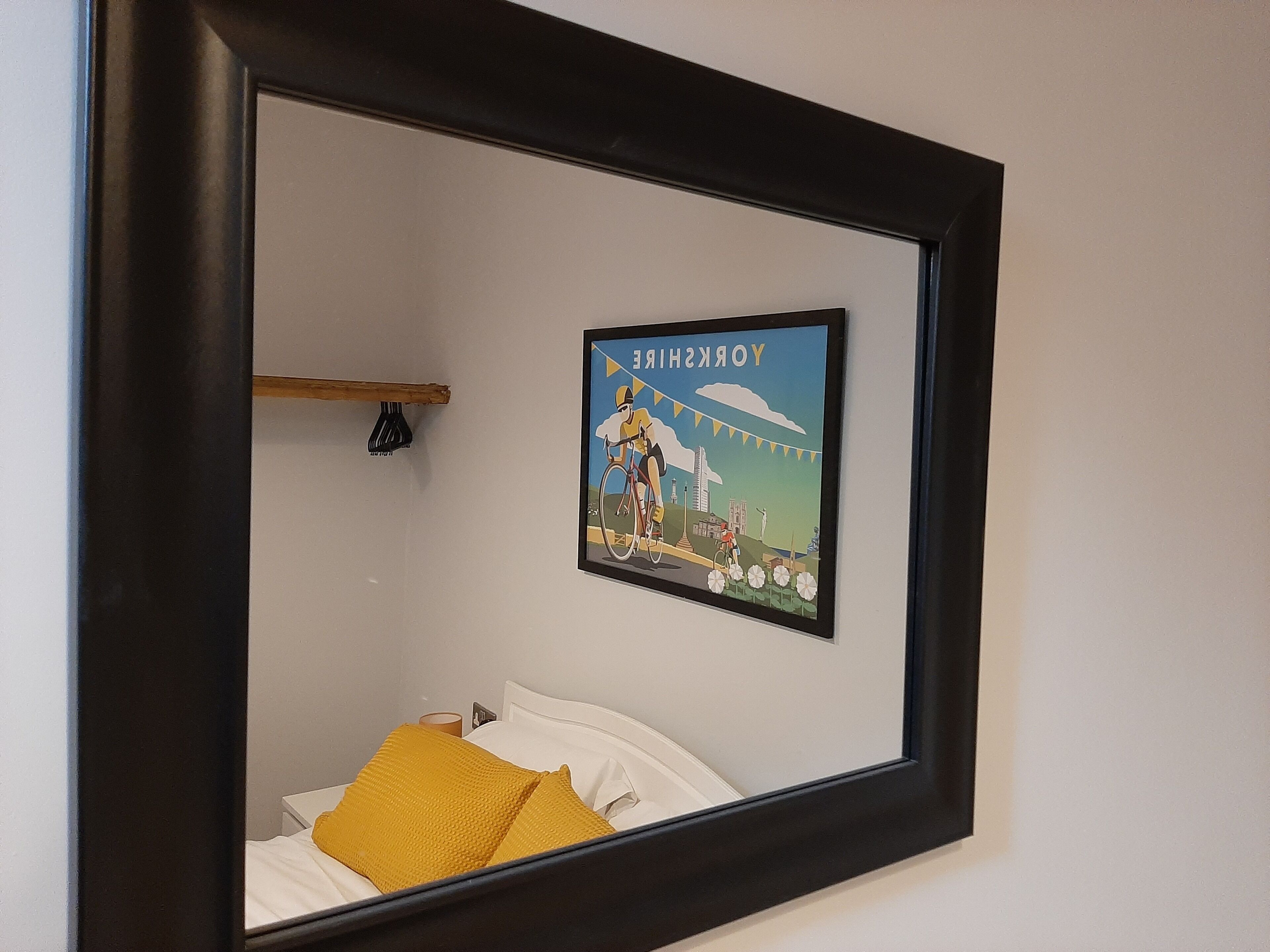Een bed met witte beddengoed en een geel kussen, een lijst met een stadsgezicht aan de muur, en een klein plankje met een zwart voorwerp.