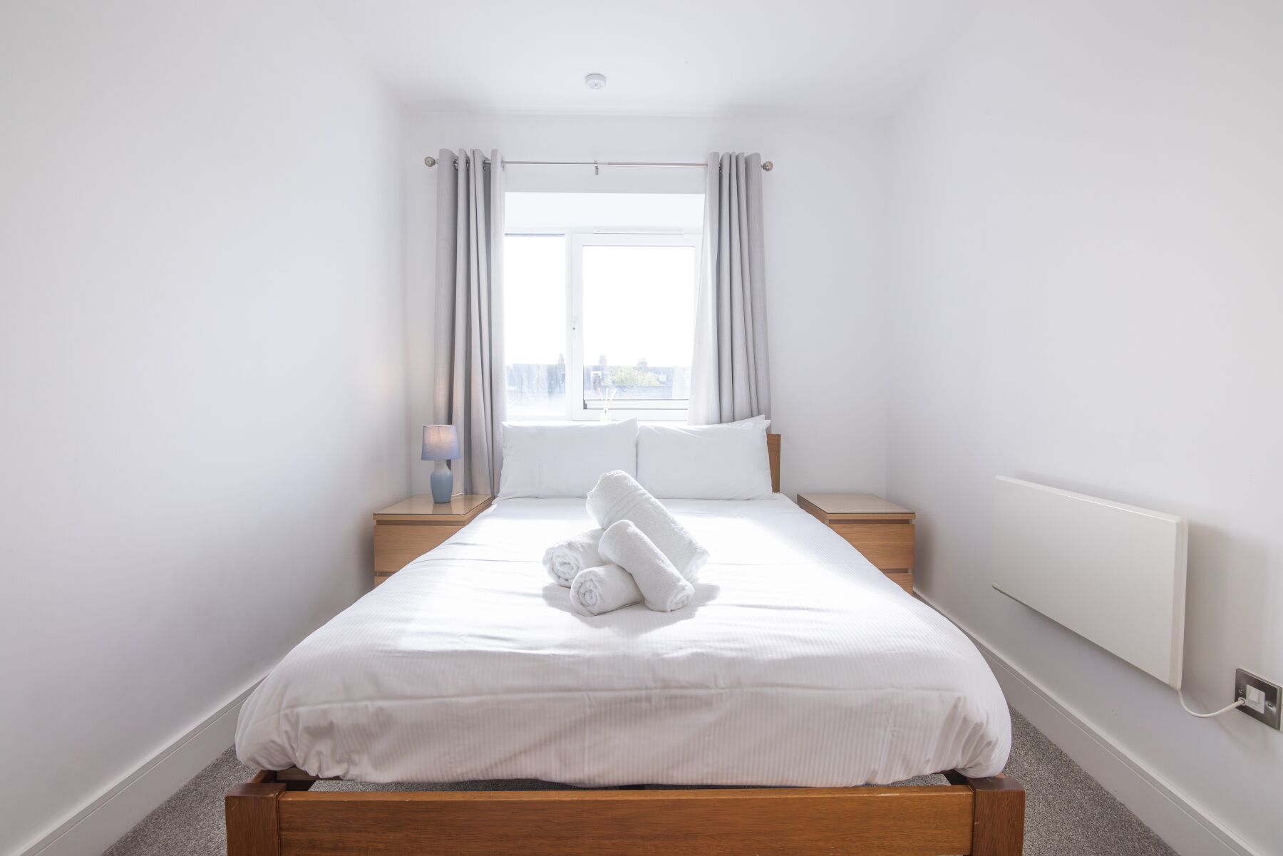 A minimalist hotel room with a single bed, a bedside table, a lamp, and a window with curtains.