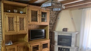 A rustic room with wooden cabinets, a flat-screen TV, a stone fireplace, and a hanging light fixture. - Casa Rural (La Horcajada)