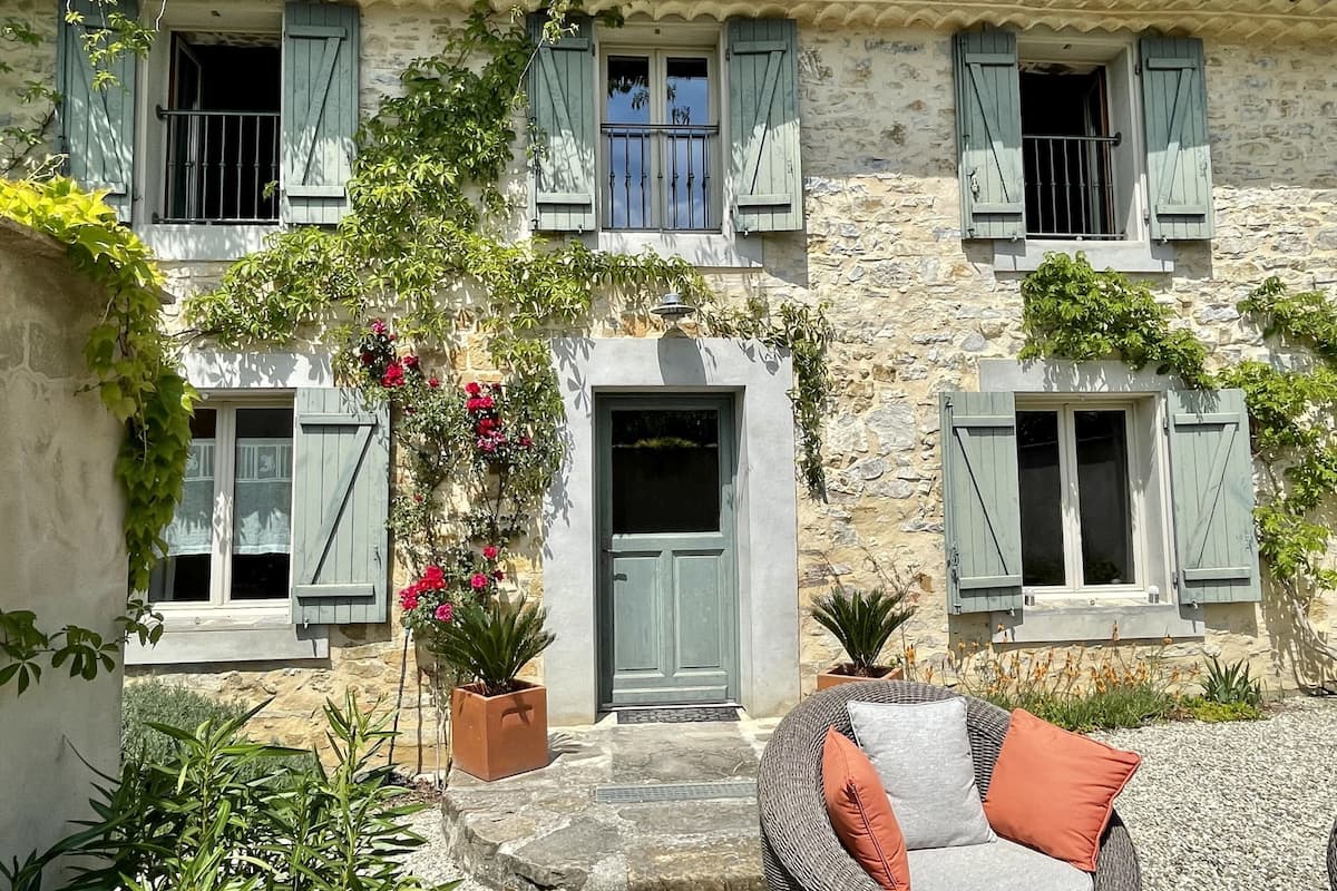 Ein Steinhaus mit einem zentralen Eingang, mehreren Fenstern mit Fensterläden und einer kleinen Veranda mit Sitzgelegenheiten.