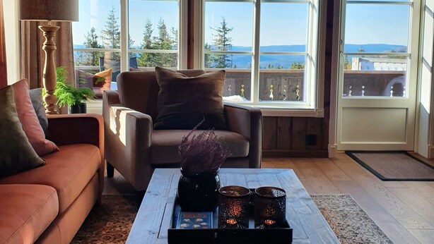 A cozy living room with a wooden ceiling, a brown sofa, a coffee table with decorative items, and large windows offering a view of trees and mountains. - Ski-in/ski out at Lifjell . Perfect Place for Hikes in the Mountains (Vestfold og Telemark)