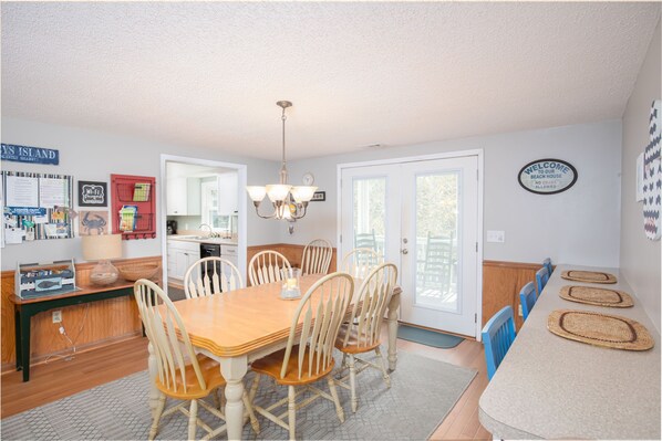 A dining area with a wooden table and chairs, a chandelier, and a kitchen in the background.