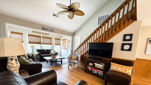 A spacious living room with a wooden staircase, leather sofas, a flat-screen TV, and a bookshelf with board games. - Beach Breezes - Oceanside, 4 bedrooms (Pawleys Island)