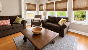 A living room with a brown sofa, a patterned armchair, a wooden coffee table, and a large window with a view of greenery. - Beach Breezes - Oceanside, 4 bedrooms (Pawleys Island)