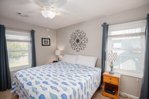 A bedroom with a bed, a ceiling fan, a wall decoration, a bedside table with a lamp, and a window with curtains.