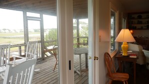 Une terrasse en bois avec des chaises et une table, une porte donnant sur une pièce où se trouvent une lampe et des livres, et une vue sur un paysage verdoyant.