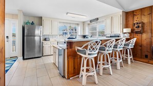 A kitchen with white cabinets, a stainless steel refrigerator, a wooden island with bar stools, and a view of the outside. - Allston - 4 bedrooms, Oceanfront (Pawleys Island)