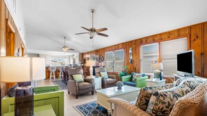 A spacious living room with a green sofa, a glass coffee table, and a television. There is a ceiling fan, a kitchen in the background, and a dining area with white chairs. - Allston - 4 bedrooms, Oceanfront (Pawleys Island)