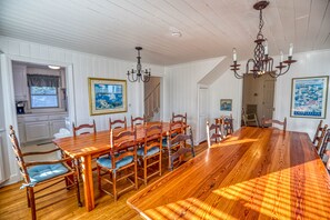 A dining room with a large wooden table and matching chairs, white paneled walls, and a chandelier.
