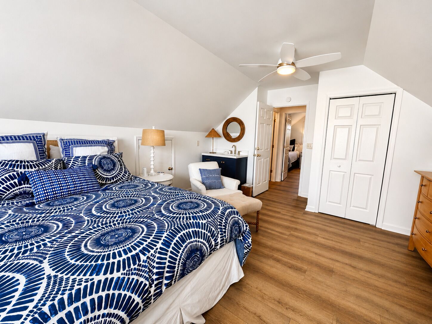 Une chambre à coucher comprenant un lit, des tables de chevet, une lampe, un ventilateur de plafond, une commode et une porte.