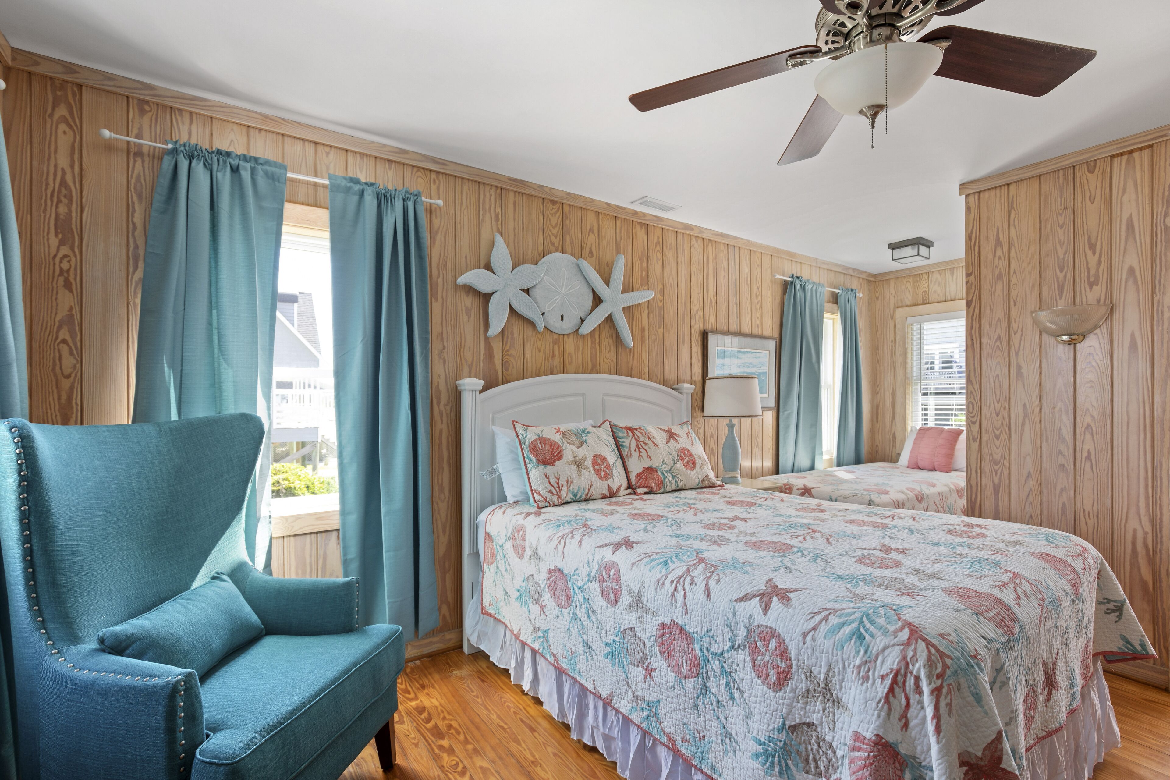 A bedroom with two beds, a ceiling fan, wooden paneling, and a large window with curtains.