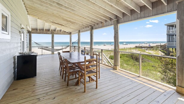 Terraza cubierta al aire libre con mesa y sillas de madera, con vista a una playa y al océano.