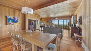 A spacious wooden interior with a large dining table, chairs, and a view of the ocean through large windows.