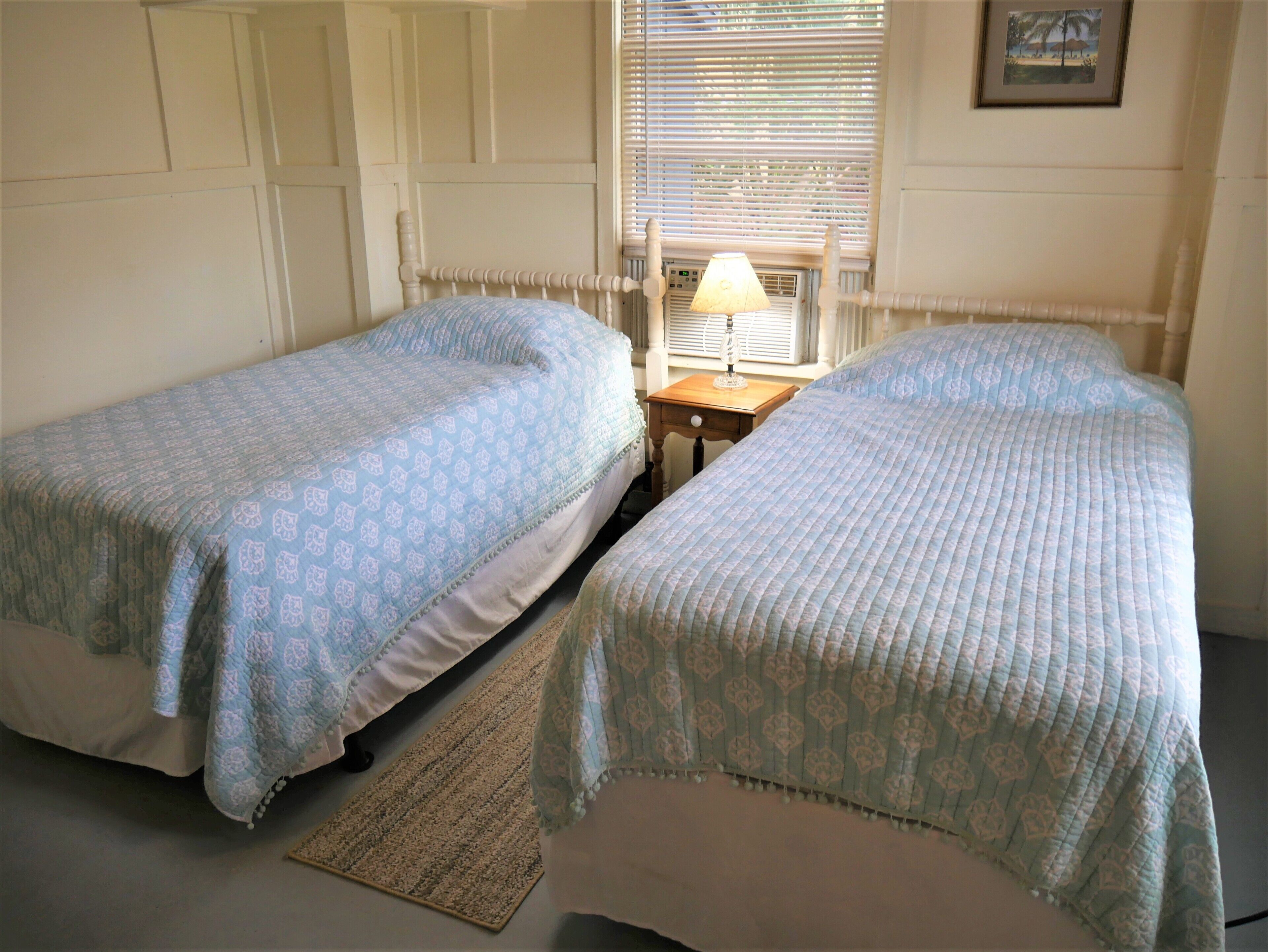 A room with two single beds, a small table with a lamp, and a window with blinds.