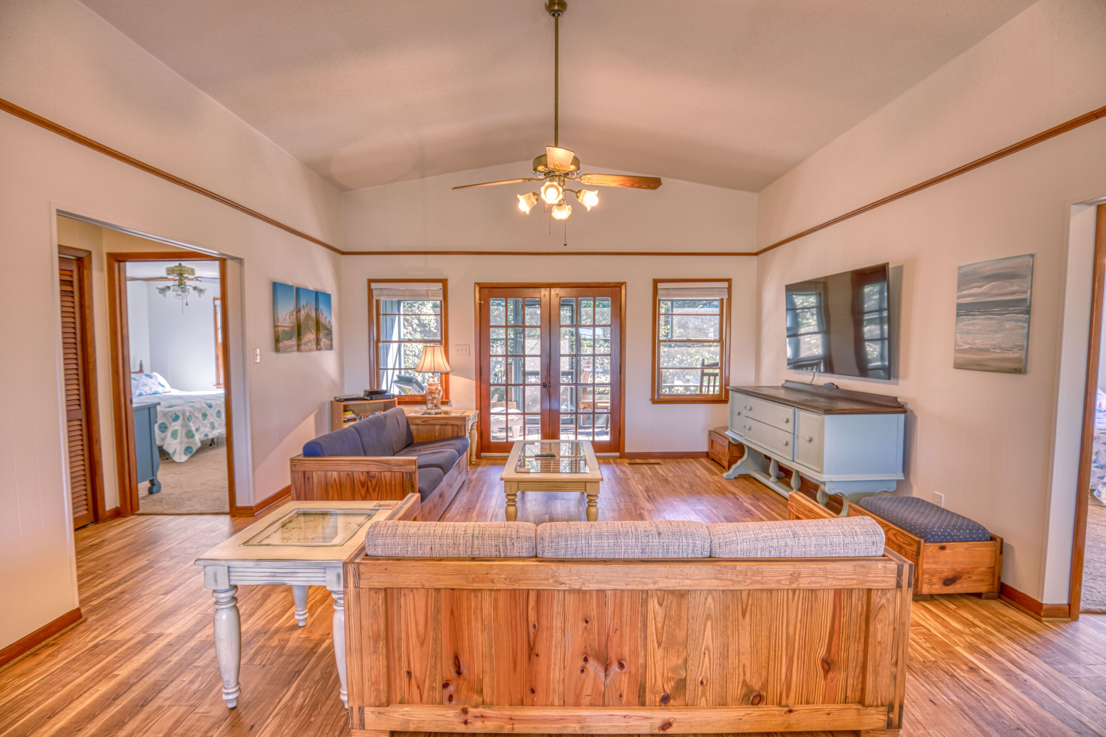 A spacious living room with a wooden sofa, a coffee table, and a ceiling fan. There are large windows and a door leading to another room.