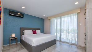 Una habitación de hotel moderna con una cama grande, una mesita de noche pequeña, un aire acondicionado instalado en la pared y un ventanal con cortinas translúcidas.
