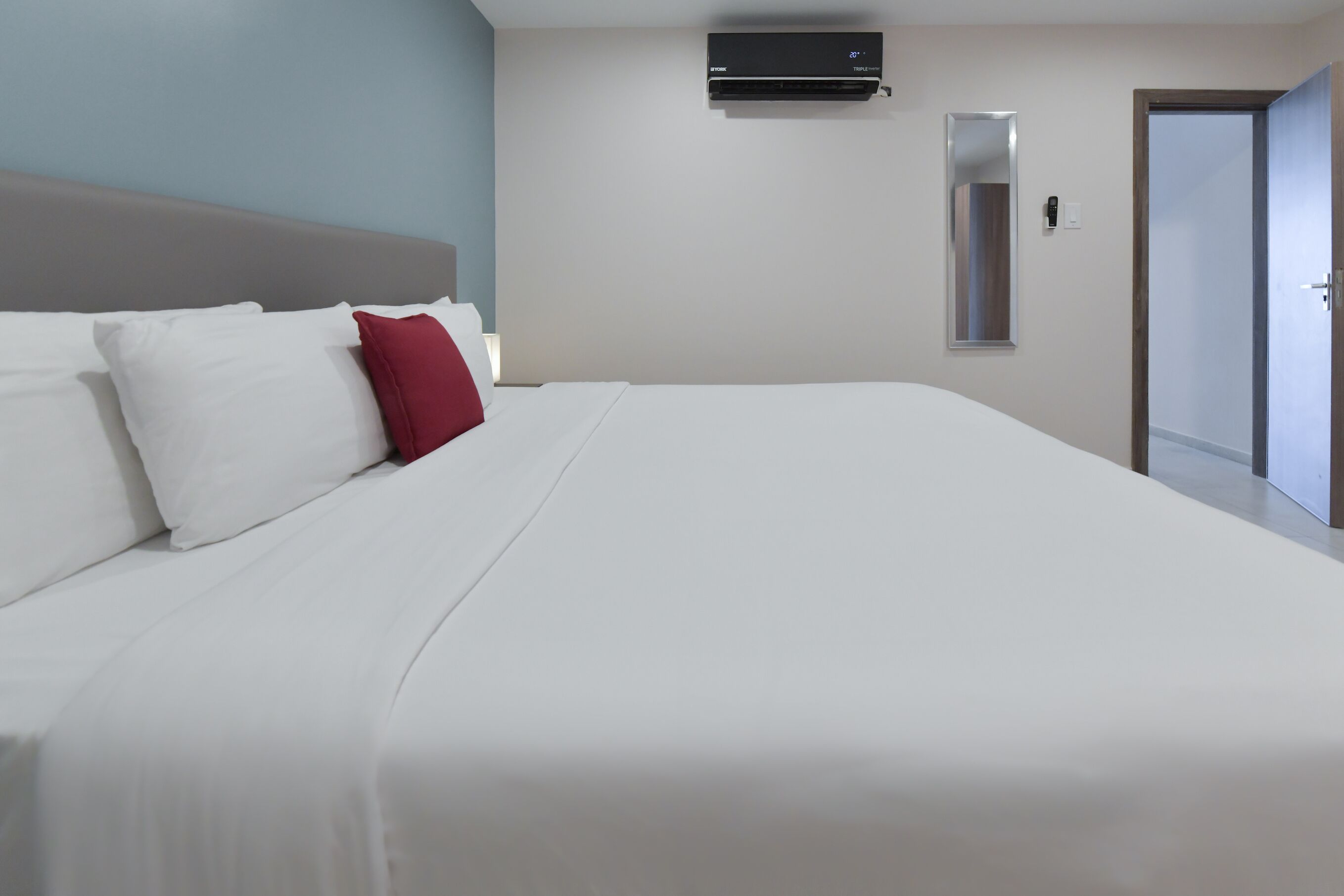 A hotel room with a large bed, white bedding, a red pillow, a wall-mounted air conditioner, a mirror, and a door.
