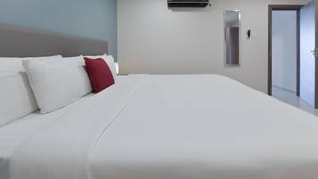 A hotel room with a large bed, white bedding, a red pillow, a wall-mounted air conditioner, a mirror, and a door.