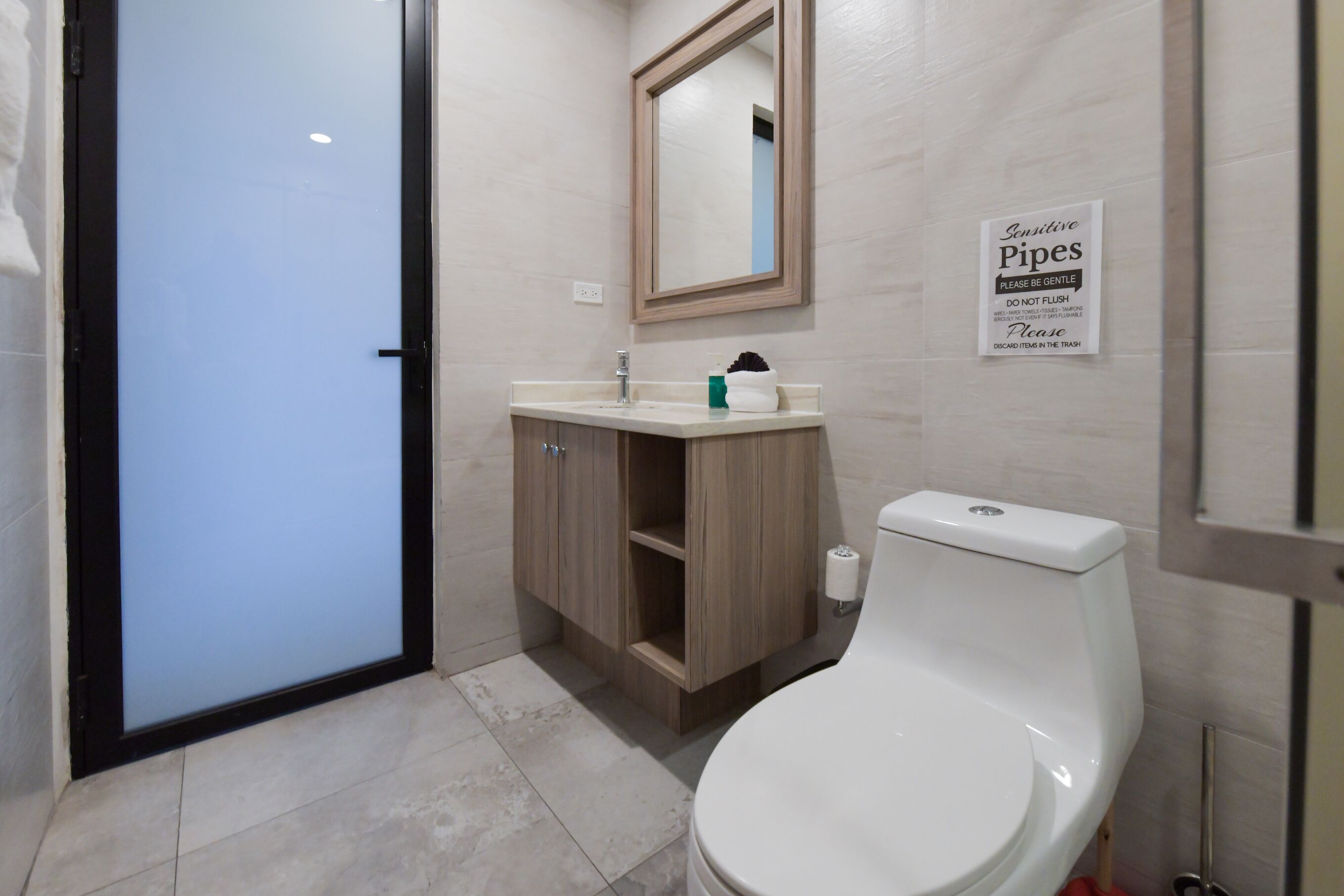 Une salle de bain avec des toilettes, un lavabo et un miroir. Un panneau est affiché sur le mur pour signaler la présence de canalisations fragiles.