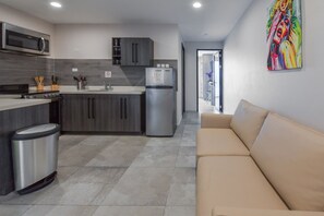A modern kitchen with grey cabinets, a stainless steel refrigerator, and a microwave. There is a beige sofa and a colorful abstract painting on the wall. - Rooftop Pool w/Lounge Area-1BE1BA Sleeps 2 #A6 (Oranjestad)