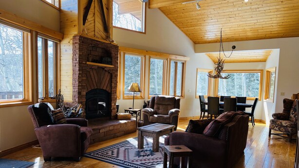A spacious living room with a fireplace, large windows, wooden floors, and a high ceiling. - Mountain Chalet Tucked in Aspen Grove with Mountain Views (Angel Fire)
