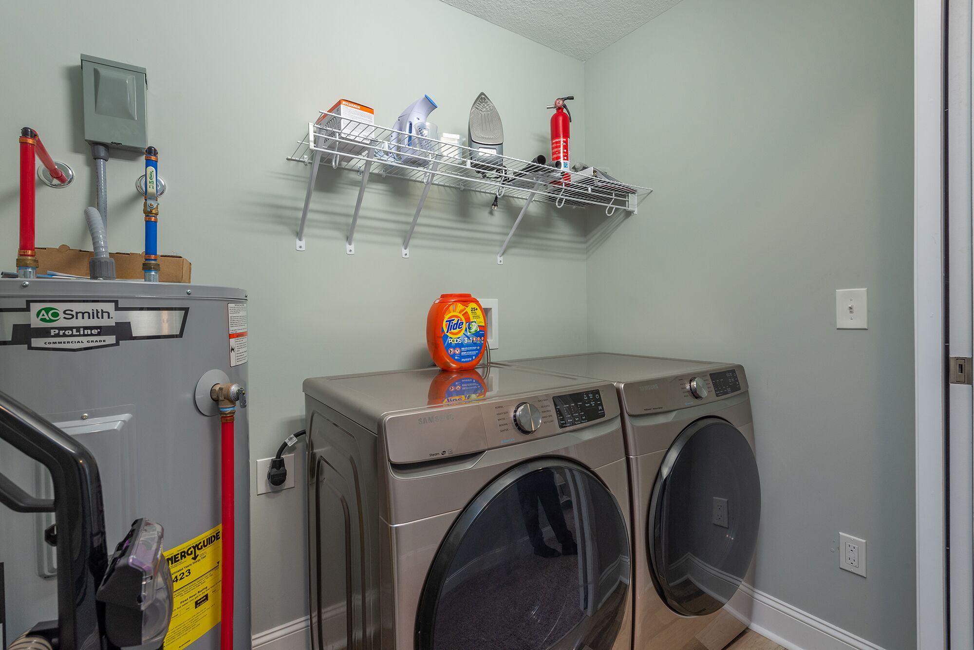 A laundry room with two front-loading washing machines, a ProLine commercial-grade dryer, and a shelf above holding cleaning supplies.