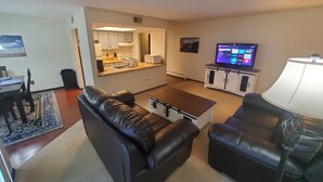 Ein Wohnzimmer mit einem schwarzen Ledersofa, einem Holztisch und einem Fernseher auf einem Schrank. Es gibt eine Küchenzeile mit weißen Schränken und einer Mikrowelle.