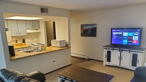 Ein modernes Wohnzimmer mit einer Ledersofa, einem Couchtisch und einem Flachbildfernseher, der an einem Schrank befestigt ist. Im Hintergrund ist eine Küchenzeile mit weißen Schränken und einer Mikrowelle zu sehen.